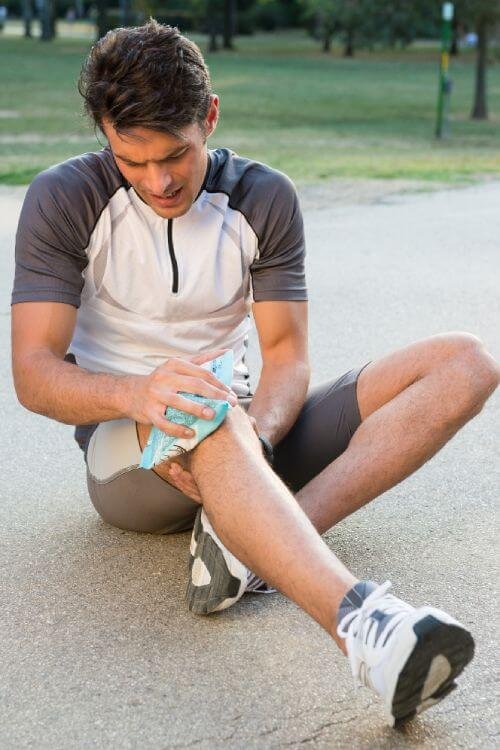 Young male athlete sitting on ground and taking ice for knee pain